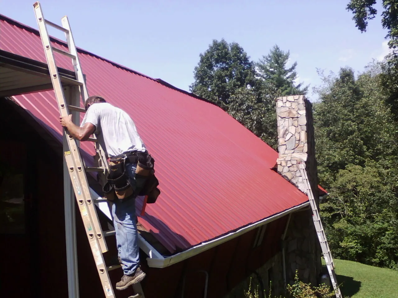 Roofer on a ladder beside a red metal roof and stone chimney — Soffit & Fascia Repair by Roofing Brothers in Molena